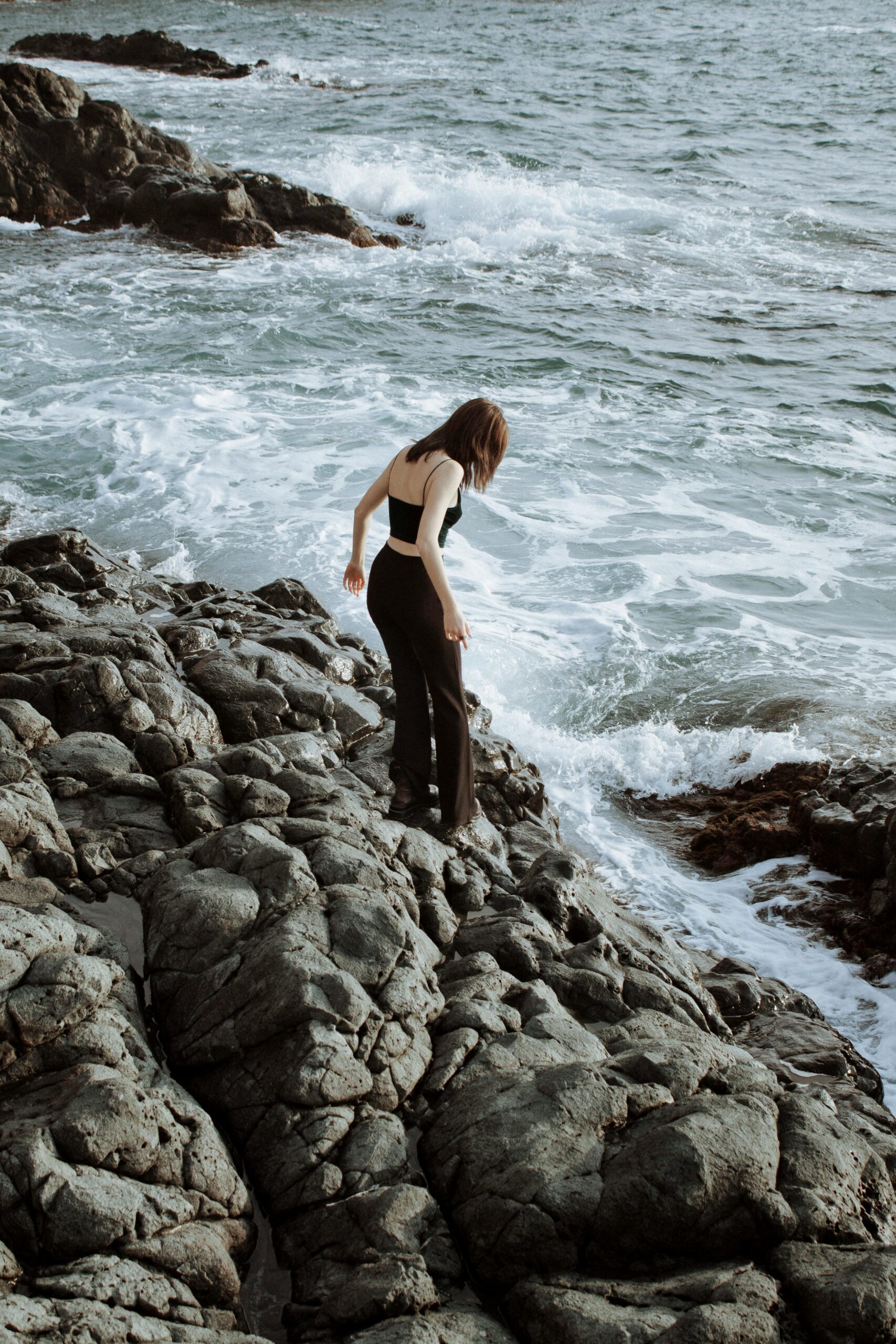 Woman exploring rocky seaside in a black outfit with ocean waves crashing nearby.