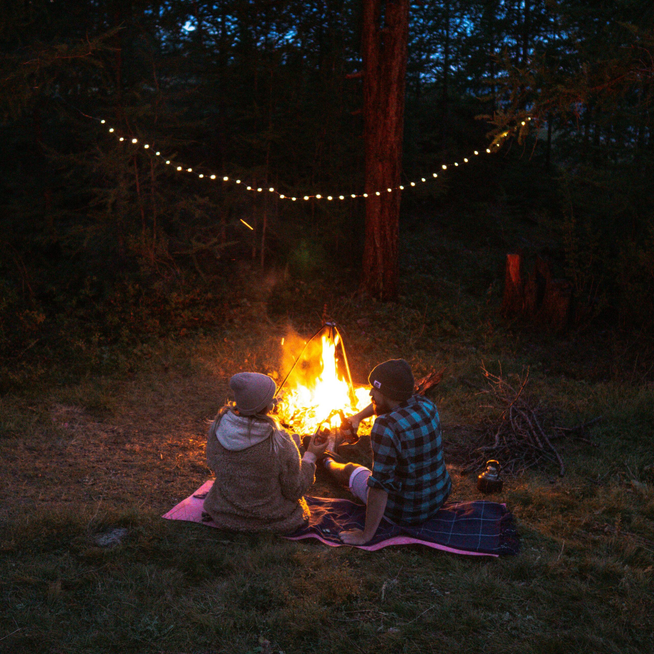 Couple enjoying a cozy evening by the campfire under string lights in a forest setting.