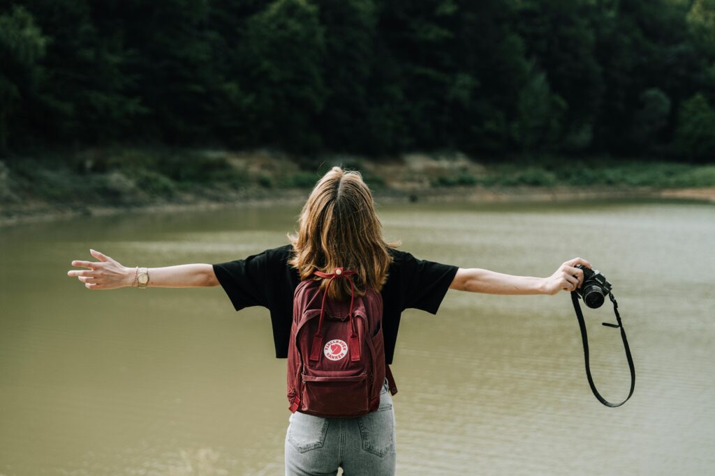 A woman stands by a lake with arms open, embracing the natural surroundings, holding a camera.