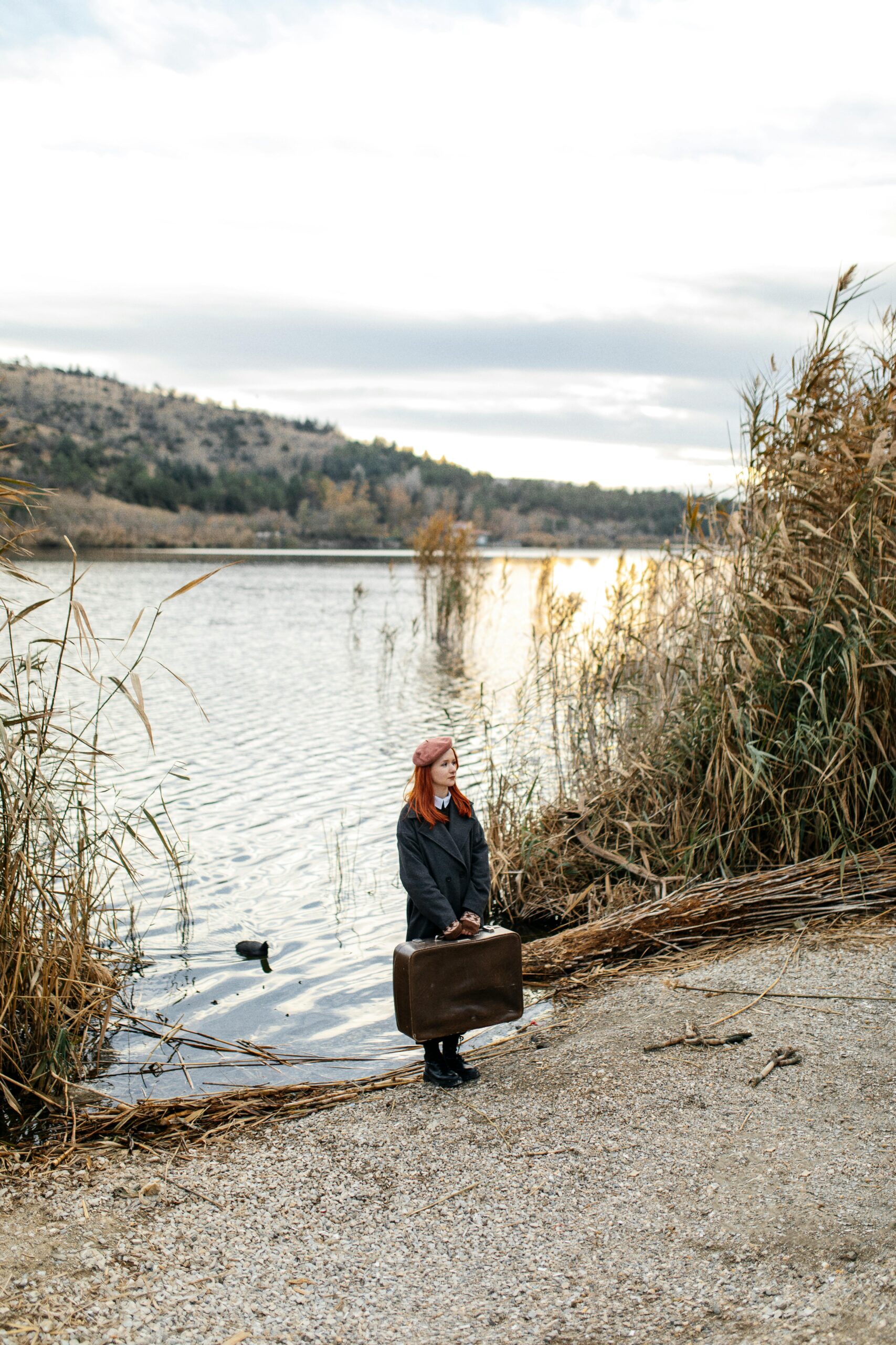 Redheaded woman standing by the river with a suitcase on a calm autumn day.