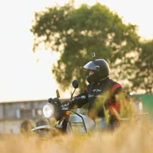 A biker riding a Royal Enfield through a sunny field under a clear sky.