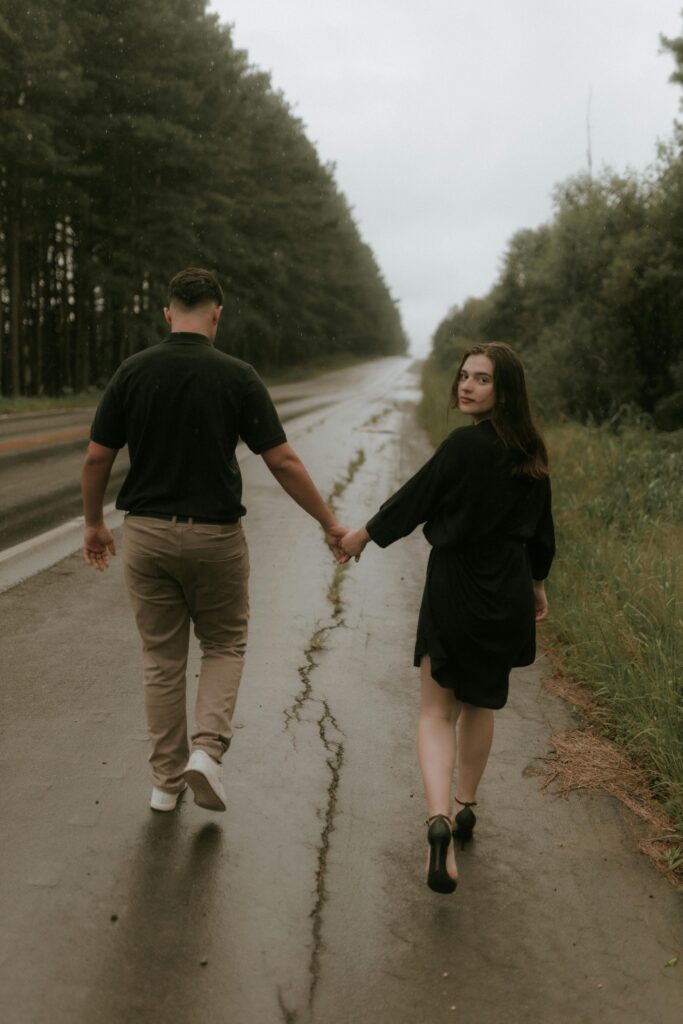 A romantic couple walks hand in hand on a scenic road surrounded by trees.