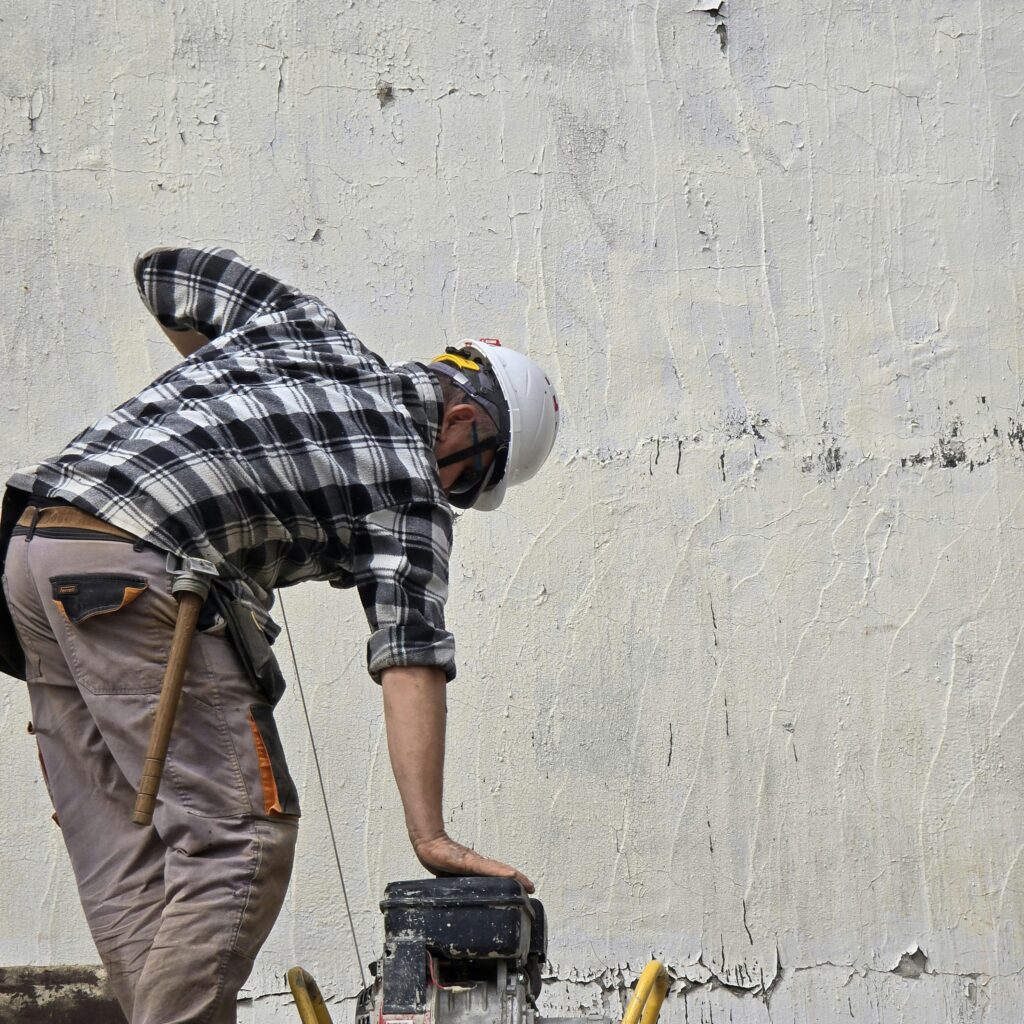 A construction worker wearing a hard hat operates machinery against a textured wall in Greece.