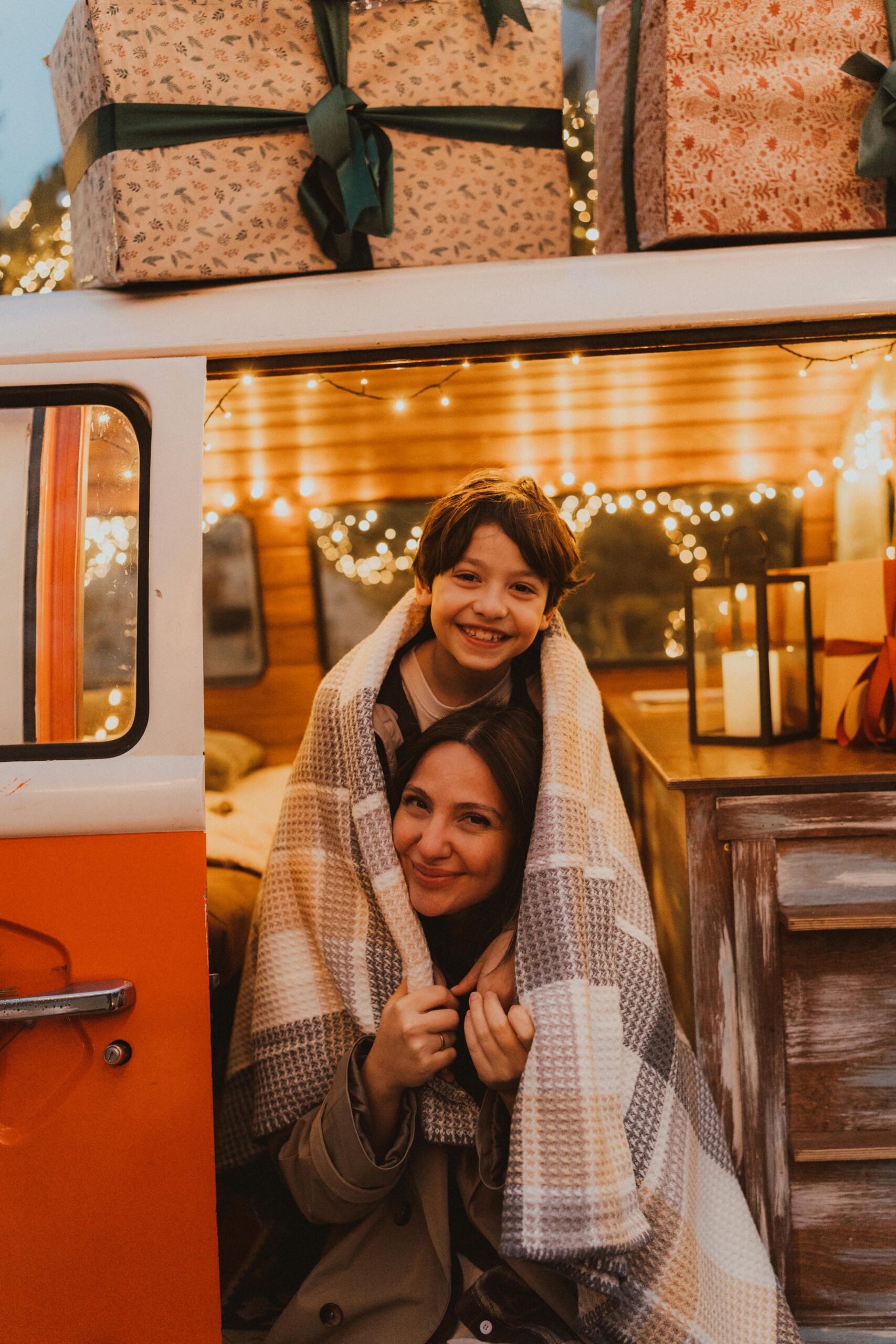 Mother and child embrace under a blanket in a decorated campervan with holiday lights and gifts.