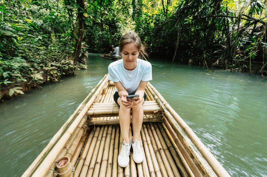 Young woman sitting on a bamboo raft in Krabi, Thailand jungle, using her phone.