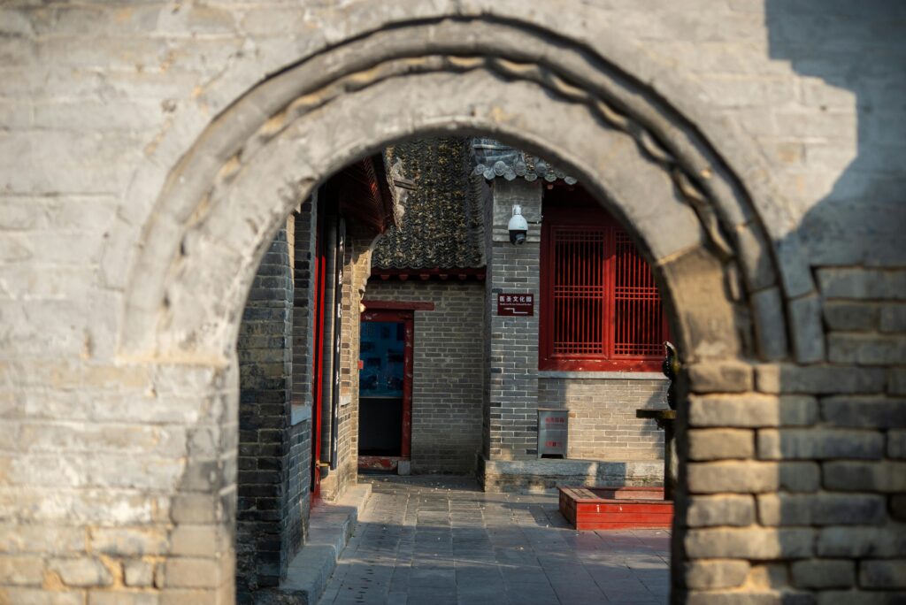 View of traditional Chinese architecture framed by a stone archway, highlighting red window shades and brickwork.
