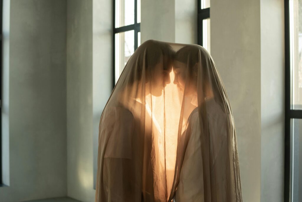 Two people in a romantic embrace under a sheer veil indoors, bathed in warm light.