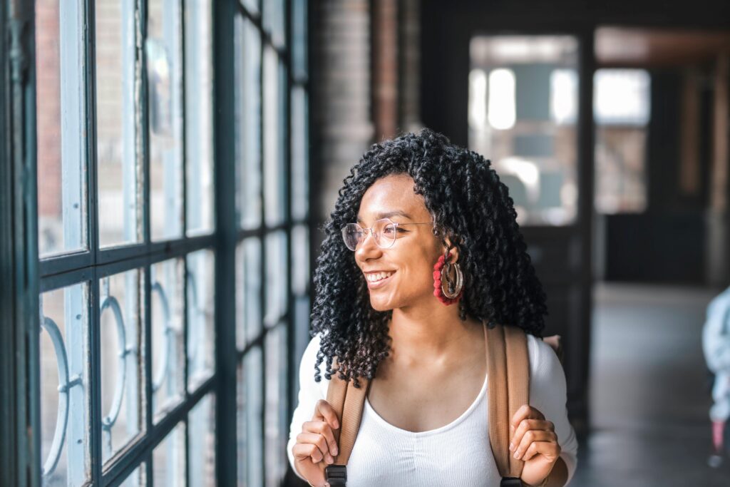 A happy woman with glasses and a backpack looks out the window in a bright corridor.