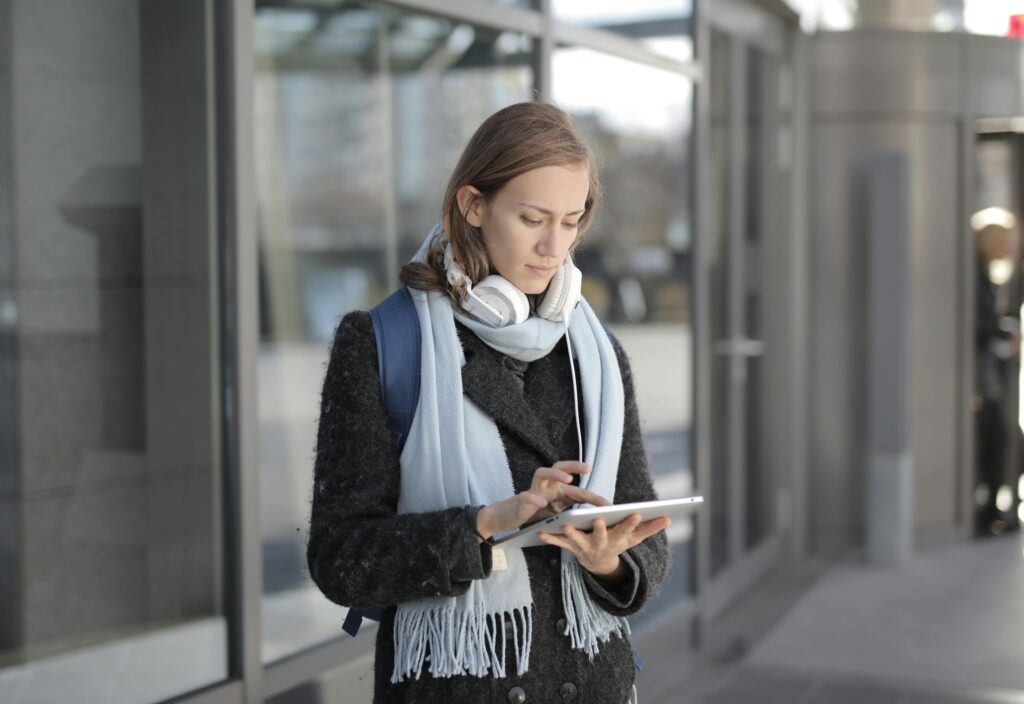 pexels-photo-3786686-3786686 Young woman stands outside using a tablet, wearing headphones and a scarf, representing technology and travel.