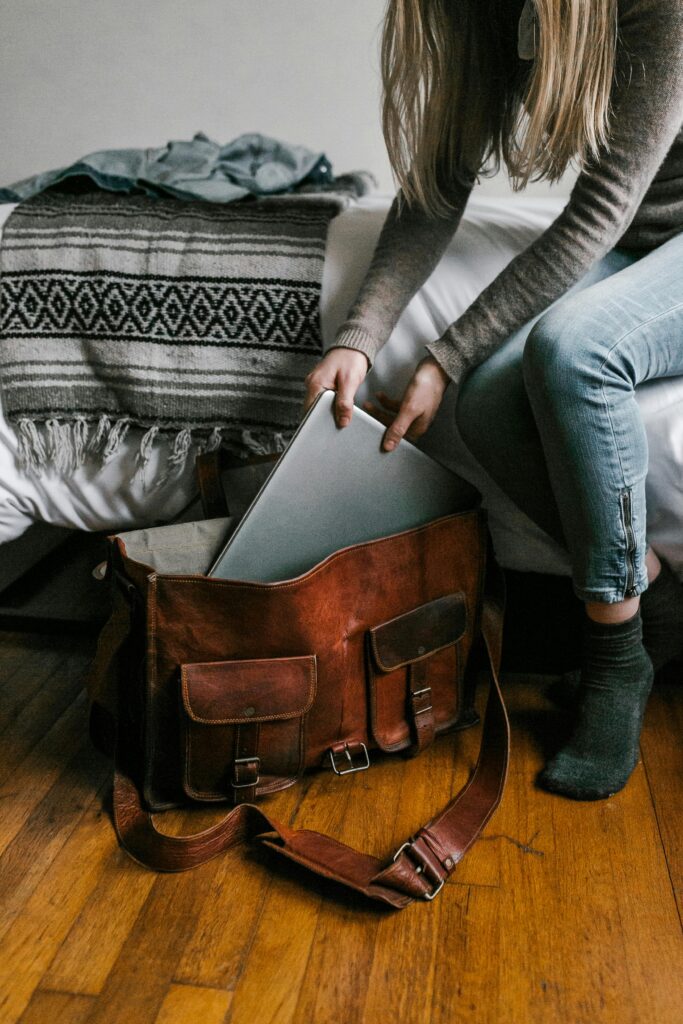 Woman packing a laptop into a stylish leather bag in a cozy bedroom setting, showcasing digital nomad lifestyle.