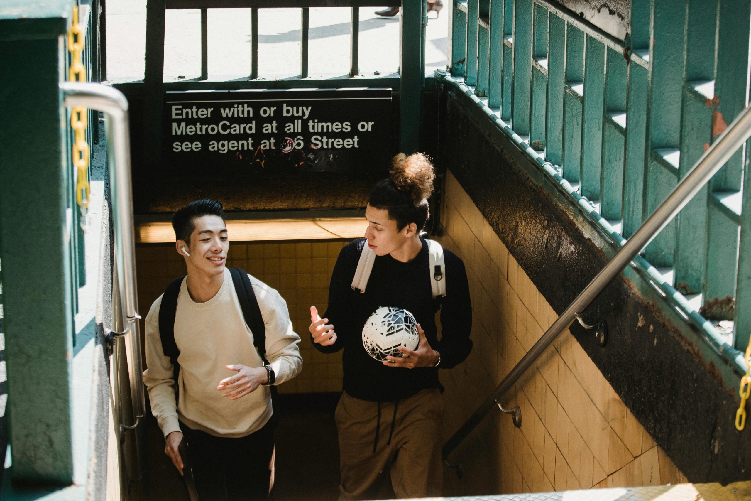 Two young men chatting as they exit a subway in New York City, one carrying a soccer ball.