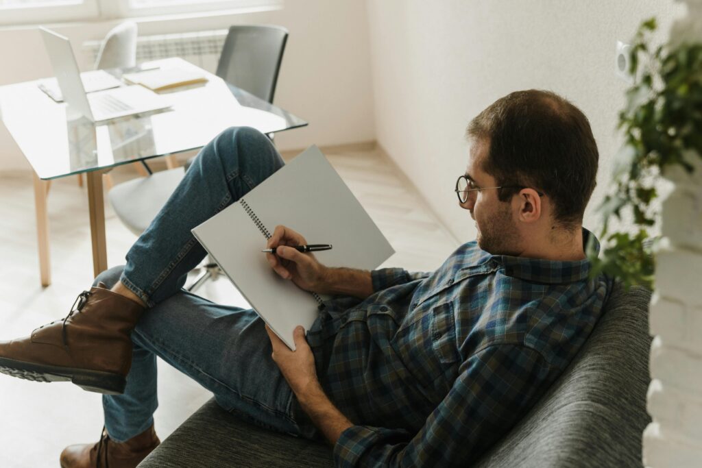 Casual man writing in a notebook while relaxing on a sofa at home.