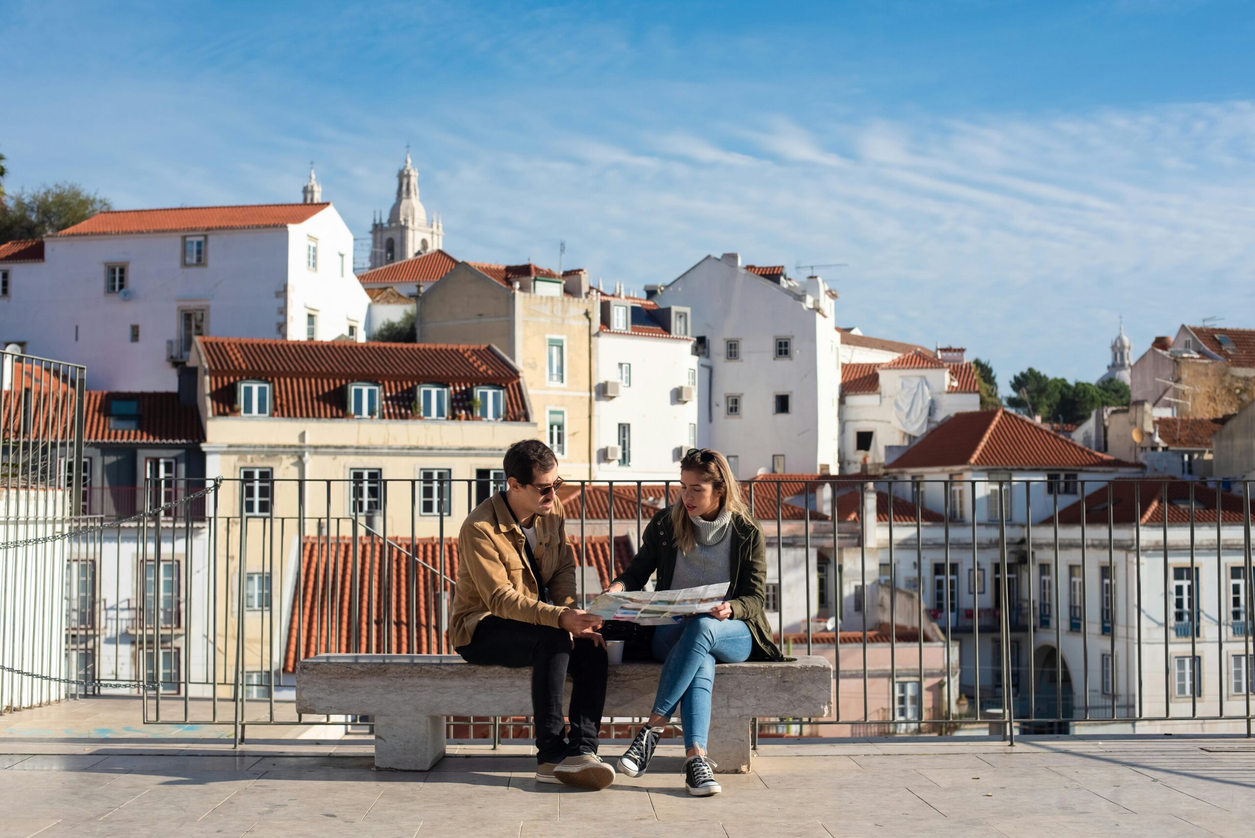 A couple sitting on a bench looking at a map with Lisbon's rooftops in the background.