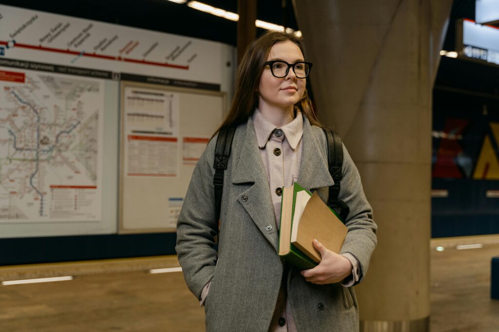 A young woman in glasses holds books while waiting at a subway station.