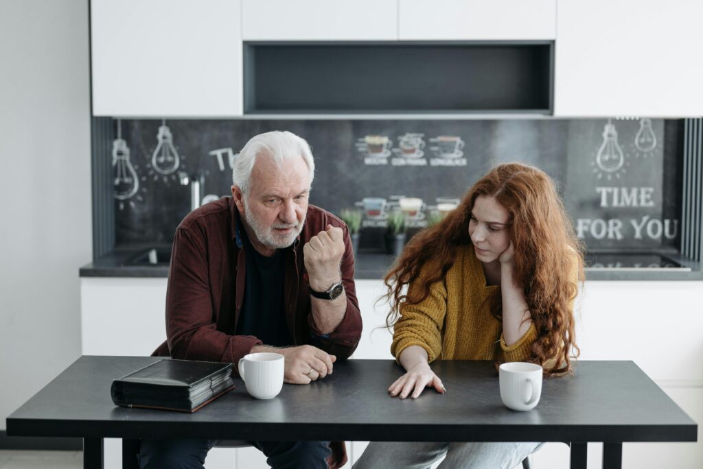 An elderly man and his granddaughter engage in a heartfelt conversation over coffee indoors.