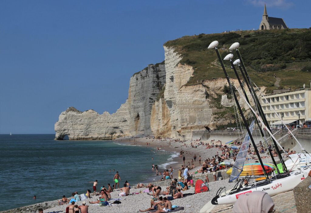 Tourists enjoy a sunny day at Étretat beach with iconic cliffs and a chapel view.