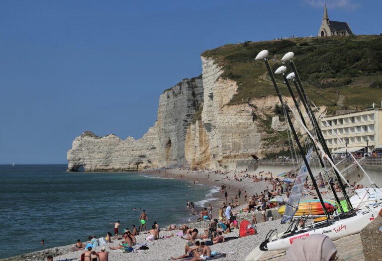 Tourists enjoy a sunny day at Étretat beach with iconic cliffs and a chapel view.