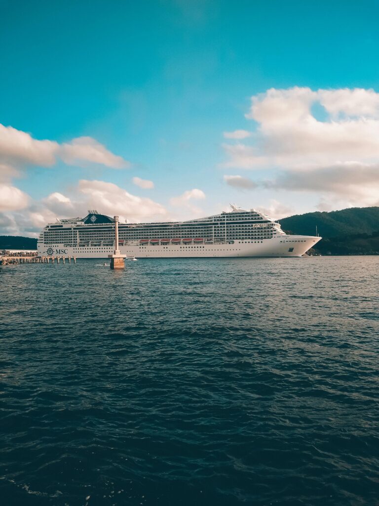 A grand cruise ship sails under a bright blue sky with fluffy clouds over a calm ocean.