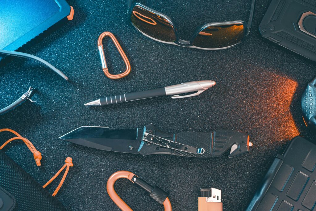 A flat lay of essential everyday carry items including a knife, pen, sunglasses, and carabiner.