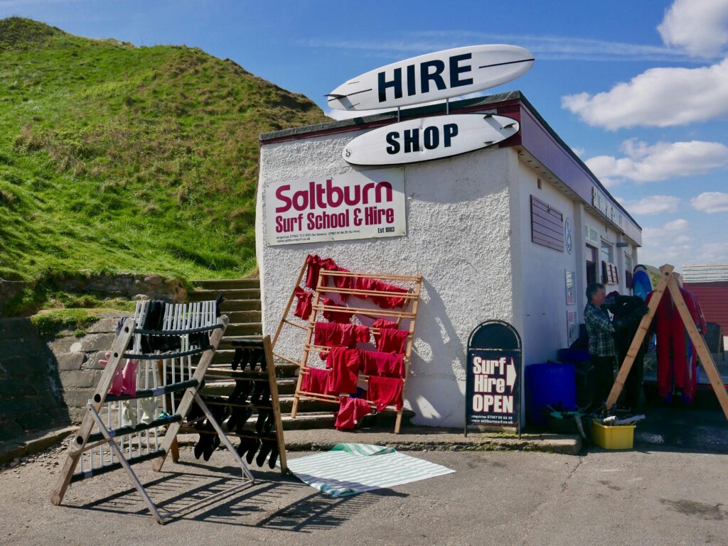 Exterior view of Saltburn Surf School & Hire shop with surfboards and gear outside under a blue sky.
