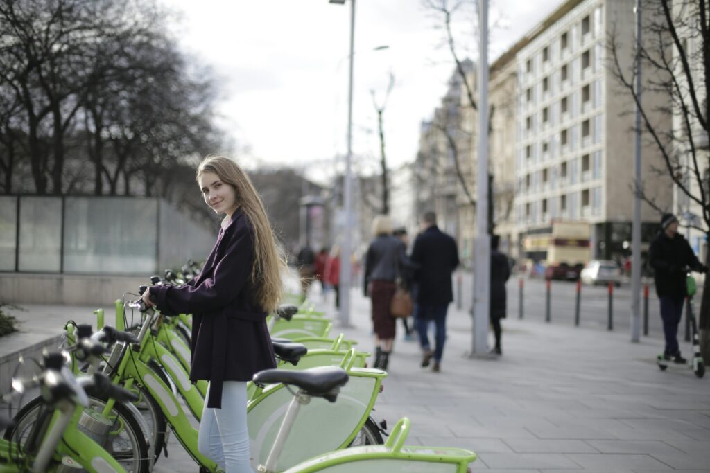 Smiling woman in violet coat by green bicycles on an urban sidewalk.
