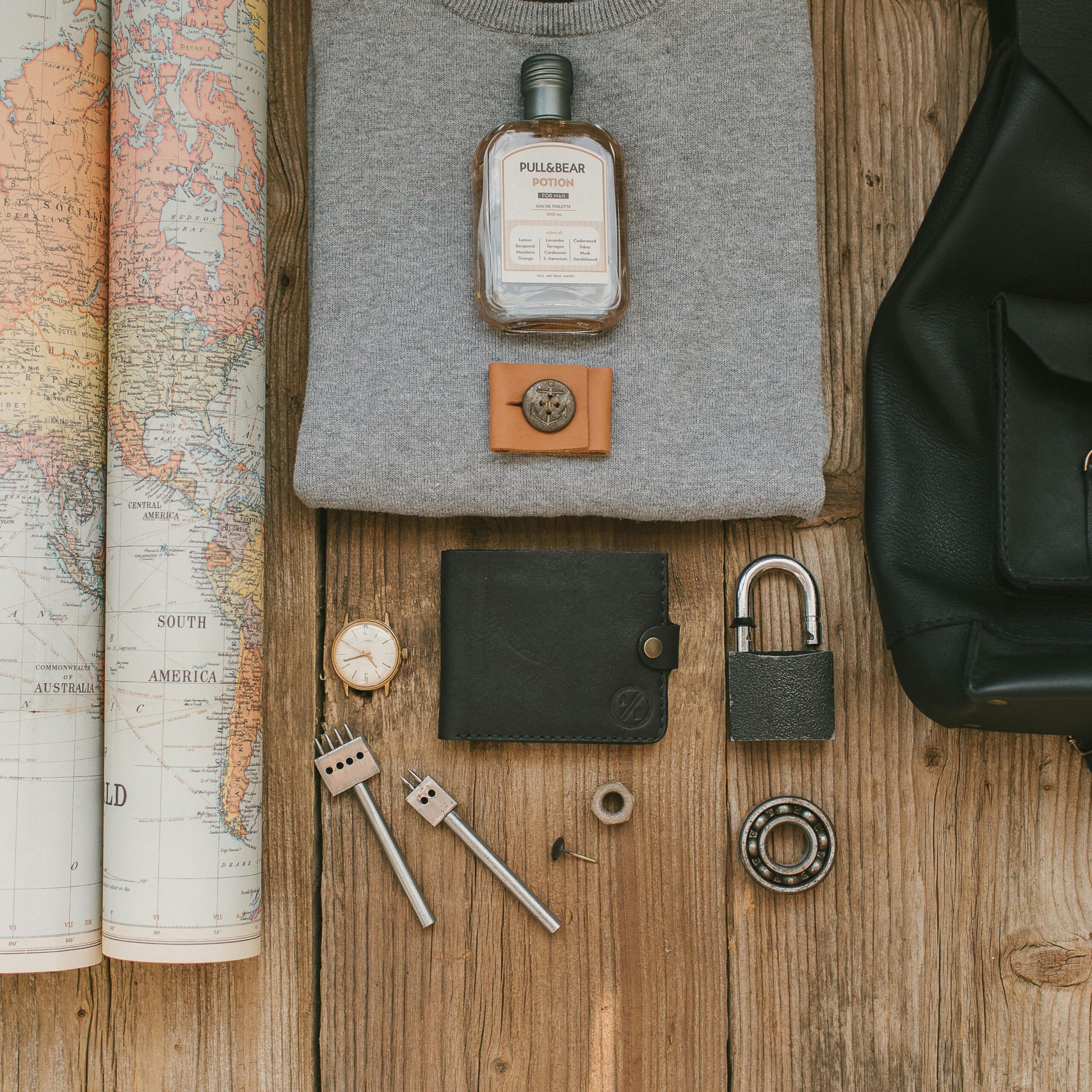 Flat lay of travel items including a map, wallet, watch, padlock, and fragrance on wooden surface.